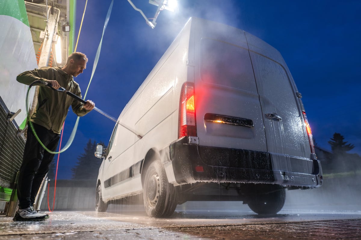 A man in a green jacket washes a white cargo van with a high-pressure hose at night
