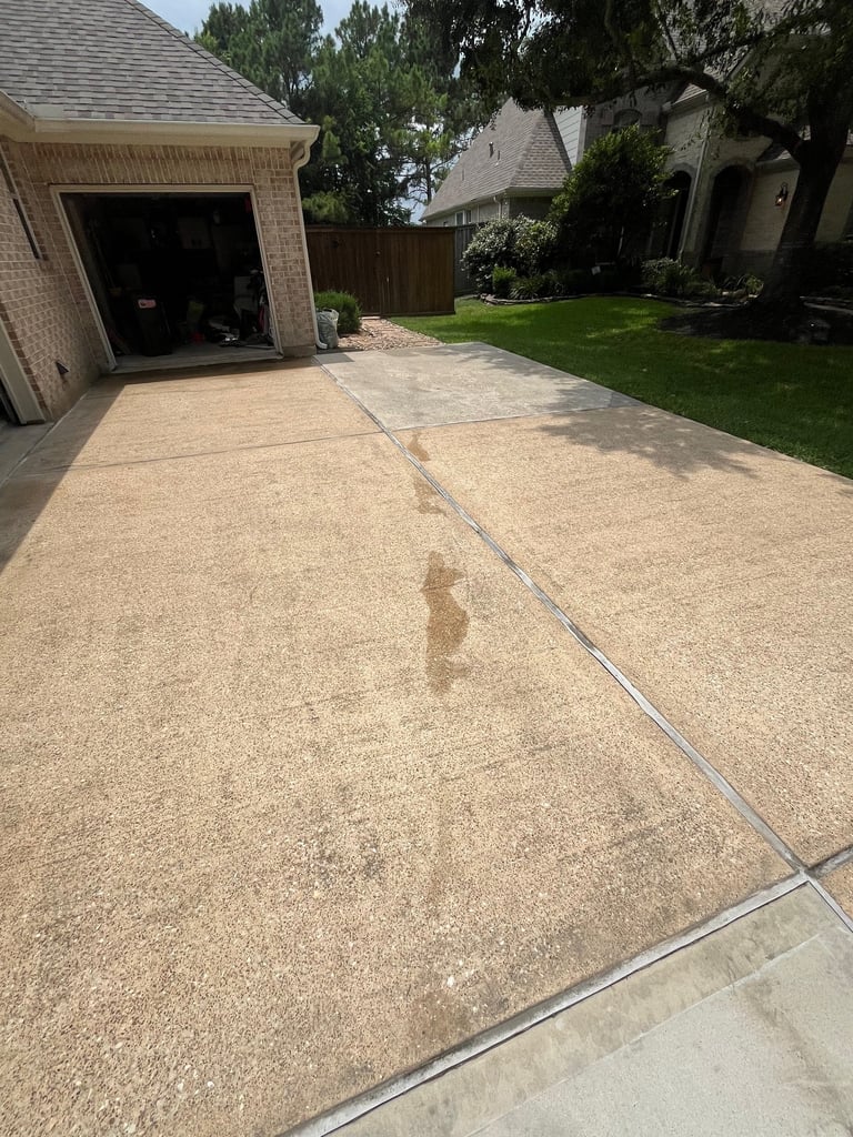 Concrete driveway with stains next to a brick garage, with lawn and trees visible in suburban residential setting