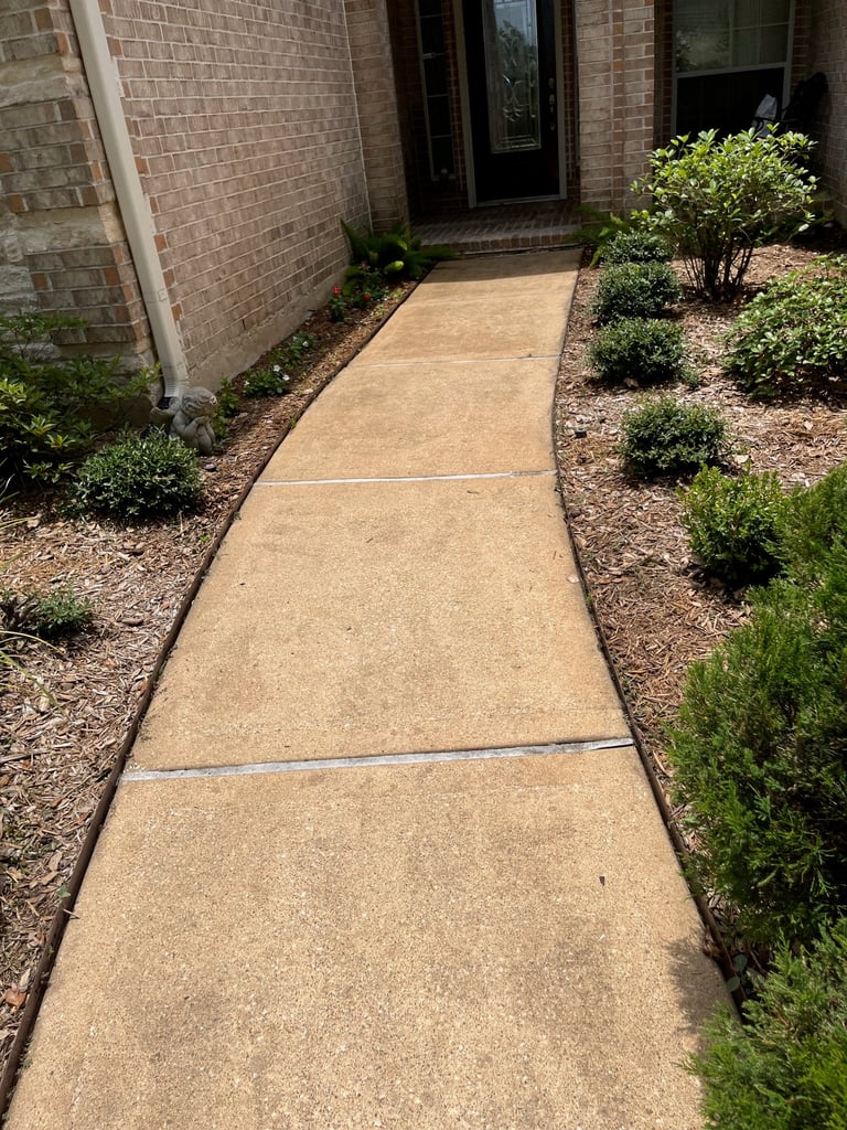 Concrete pathway leading to brick house entrance with landscaped shrubs and mulch on both sides